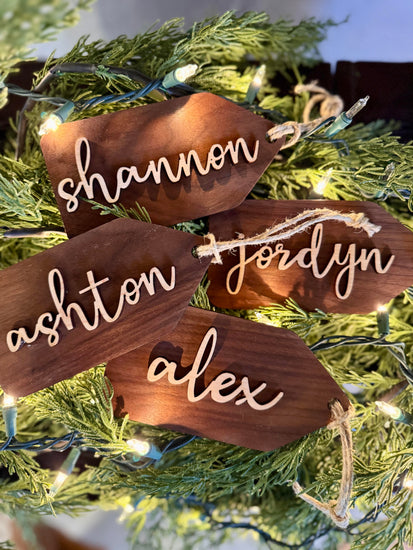 Wooden name tags/ stocking tags hanging on a Christmas tree with decorative lights.